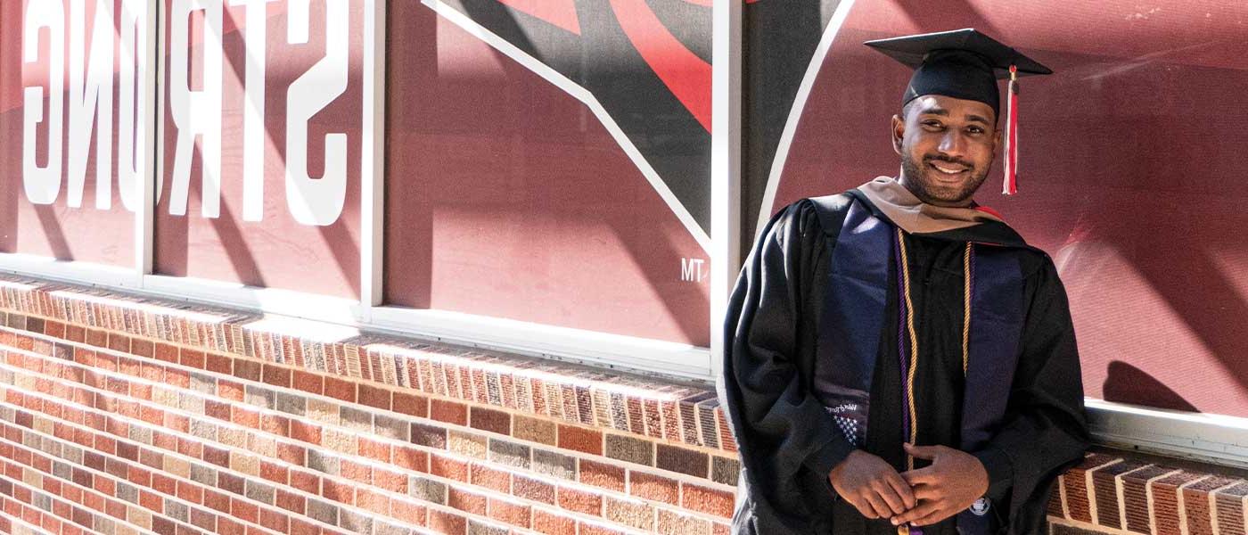 UIW veteran student in cap and graduation gown in front of the flags and chapel 